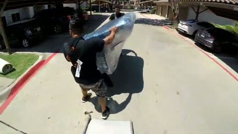 two movers carrying a wrapped sofa across an apartment complex driveway, face visible